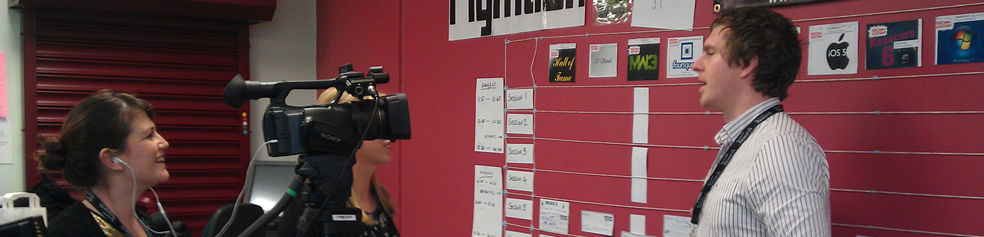 A white professor stands in front of posters while a white woman records a video of his comments